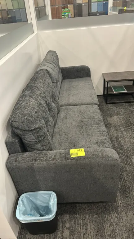 Cushioned lounge sofa. Features gray textured fabric upholstery, tufted back cushions, and two-cushion seating. 7'L. Side table and wastebasket not in