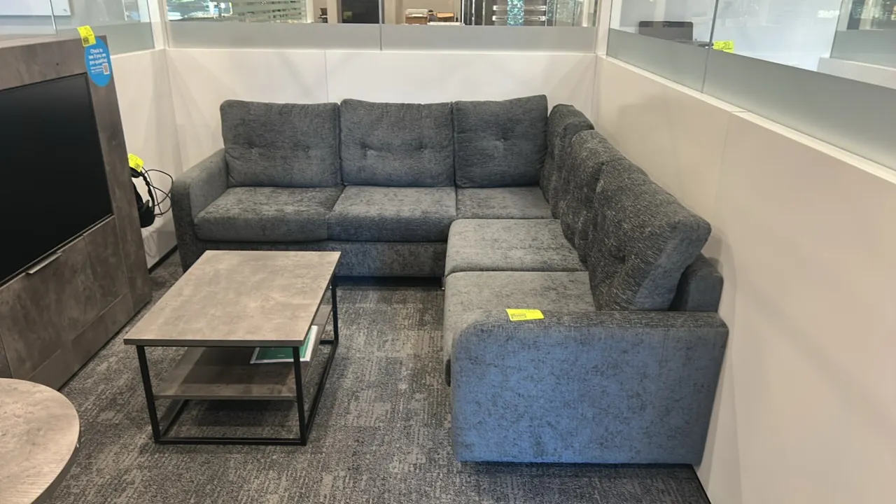 Cushioned L-shaped sectional lounge sofa. Features gray textured fabric upholstery, tufted back cushions, and chrome cylindrical feet. 7' x 7'. Coffee