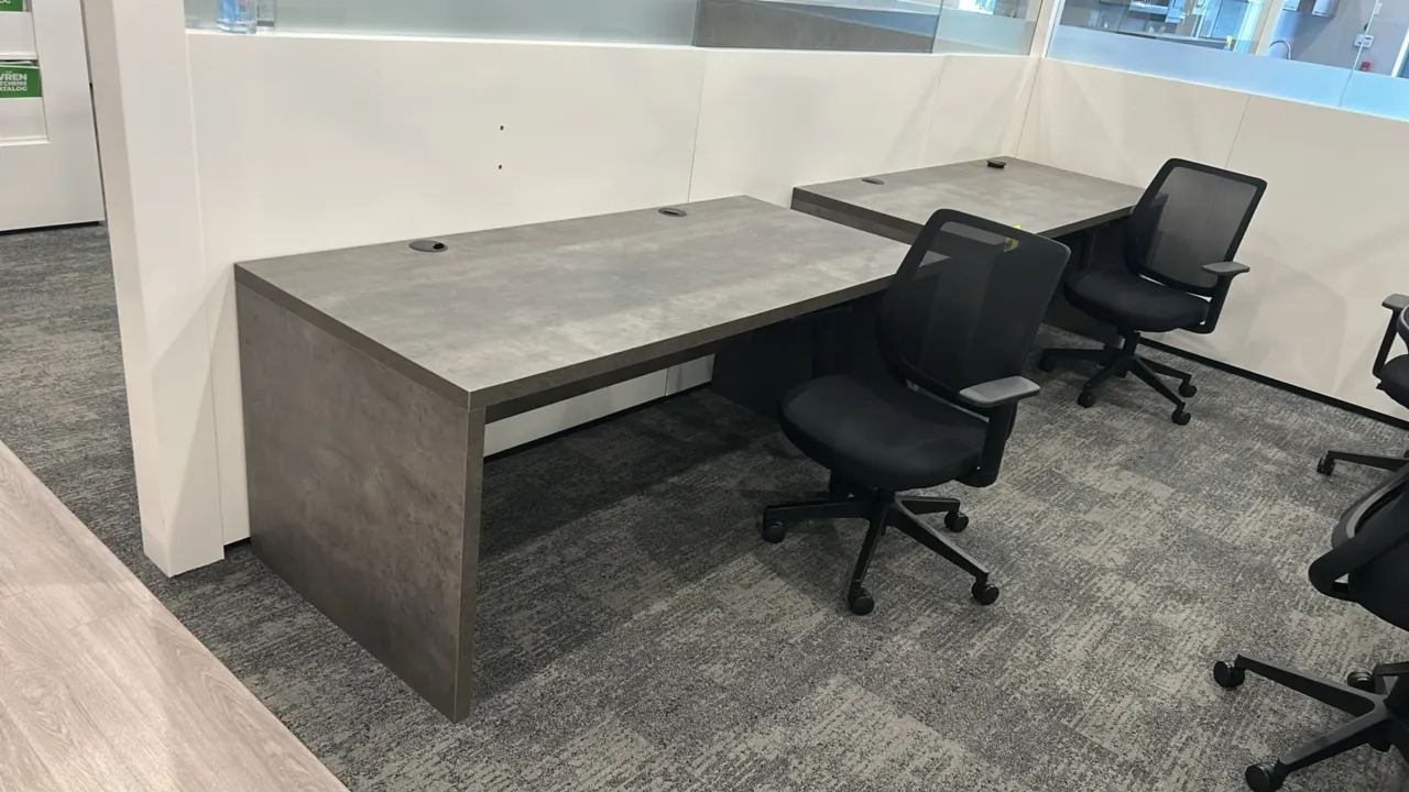 Laminate work desks with rolling office chairs. Desks feature gray textured laminate finish, panel base, and dual desktop cable pass-through grommets.