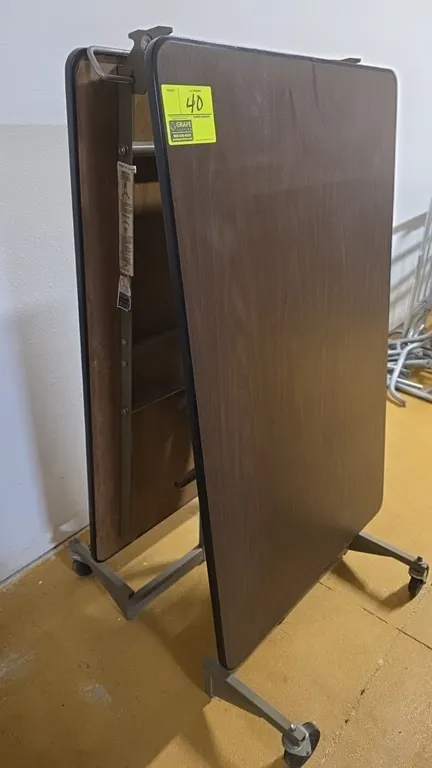KI mobile folding table. Features a woodgrain laminate top, protective edge banding, and center folding mechanism. 8'L. Mounted on a metal frame with