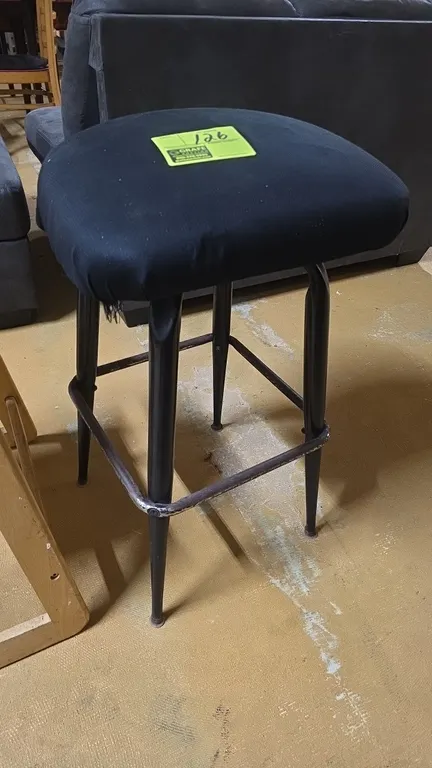 Metal stool with padded seat. Features a black fabric-covered upholstered seat, four tapered tubular metal legs, and an integrated perimeter footrest