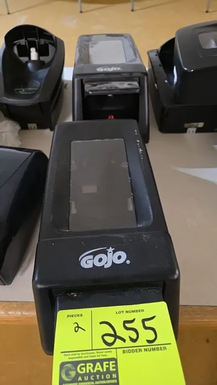 GOJO manual soap dispensers. Features black plastic construction, manual push-bar dispensing mechanism, clear sight window, and wall-mount backing.