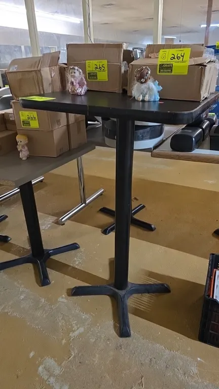 Bar-height table. Features a black laminate top with rounded corners. Dimensions: 24" x 24". Mounted on a black metal pedestal column with an X-style