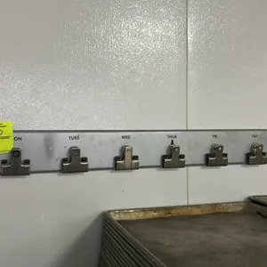 Baker's Aid metal wall-mounted organizing rack. Features 9 spring clips labeled for days of the week, hold, and special orders. Sheet pans not include