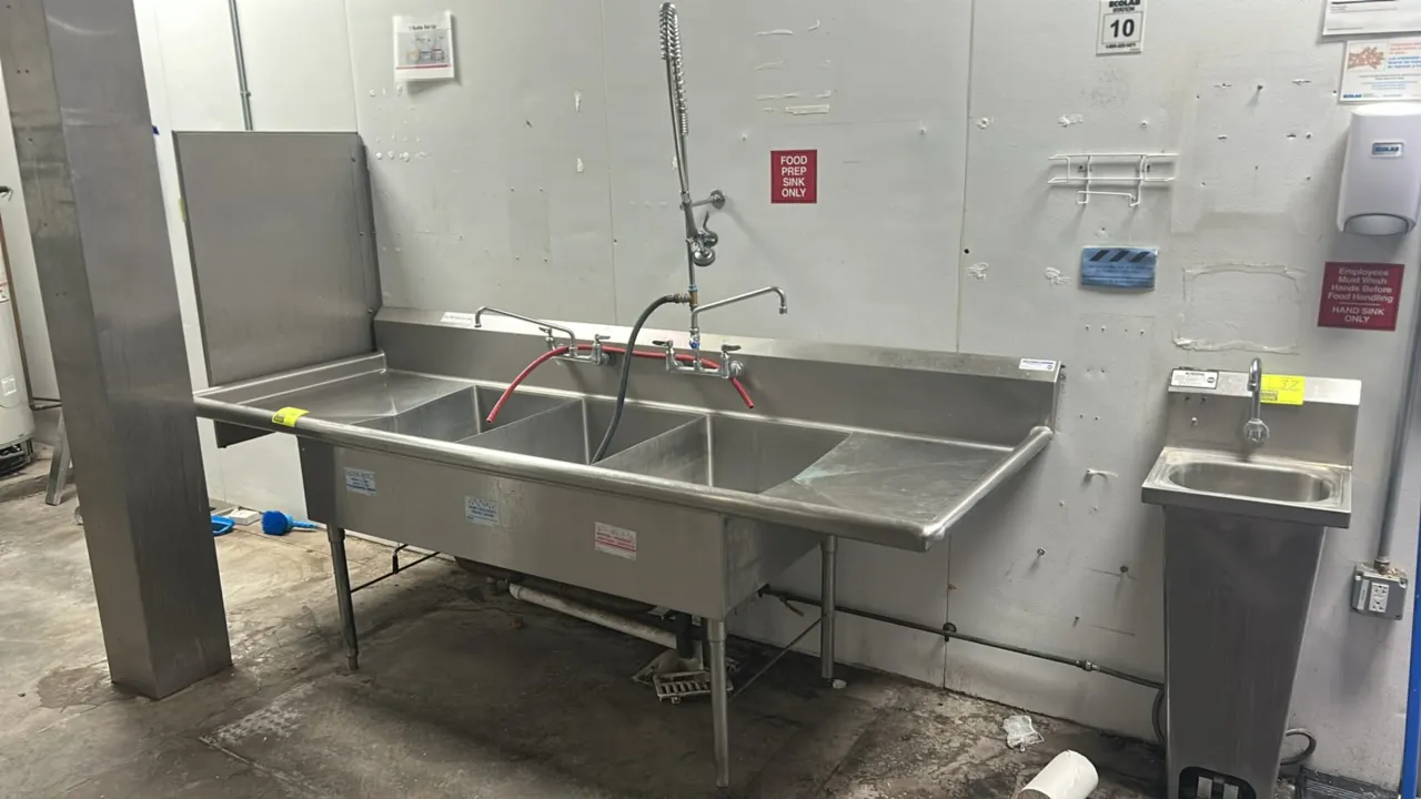 Stainless steel 3-compartment sink. Features left and right drainboards, integrated backsplash with left side splash, two splash-mount faucets, and an