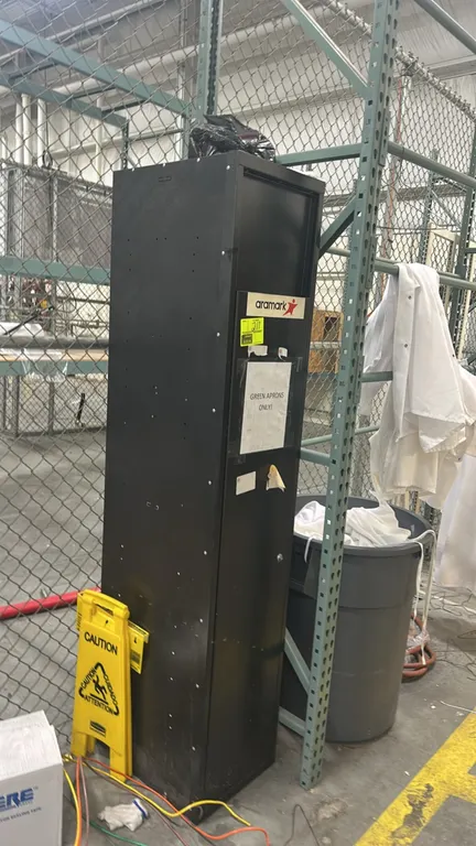 Aramark metal clothing drop-off locker. Features black metal construction, top drop door, and lower access door with key lock. Contents not included.