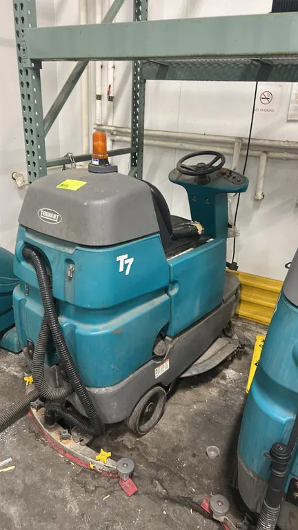 Tennant T7 micro-rider floor scrubber. Features steering wheel controls, amber warning beacon, rear squeegee assembly, and recovery hose. Note: Operat