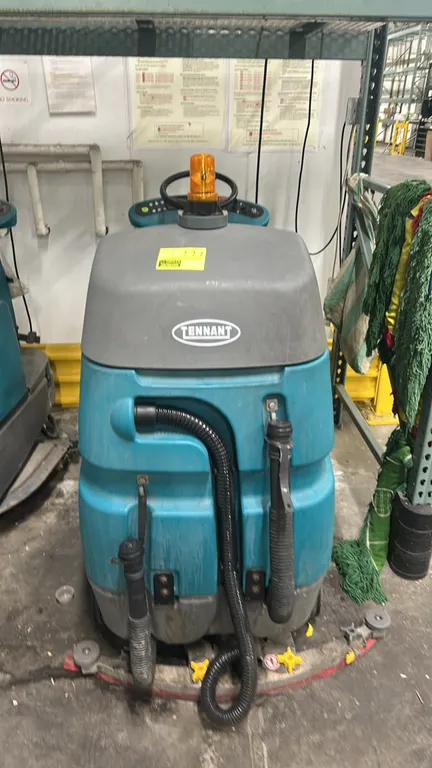Tennant T7 micro-rider floor scrubber. Features an operator seat, steering wheel controls, top-mounted amber beacon light, rear squeegee assembly, and