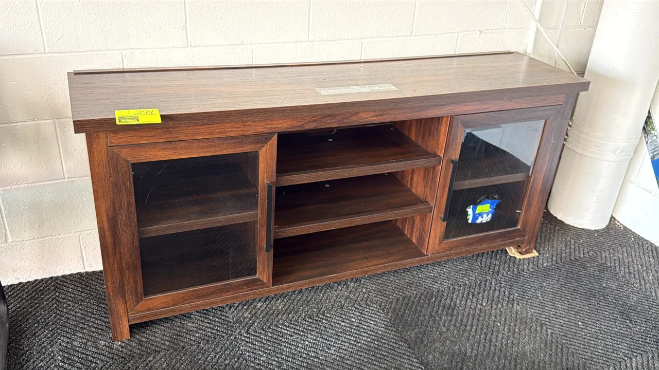 Wood laminate TV cabinet. Features woodgrain finish, two glass-paneled doors with black hardware, and open center shelving. 65"L. Contents not include