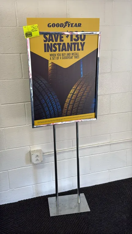 Metal sign stand. Features a chrome-plated finish, top-loading sign frame, and dual upright posts. Mounted on a flat rectangular metal base. Includes