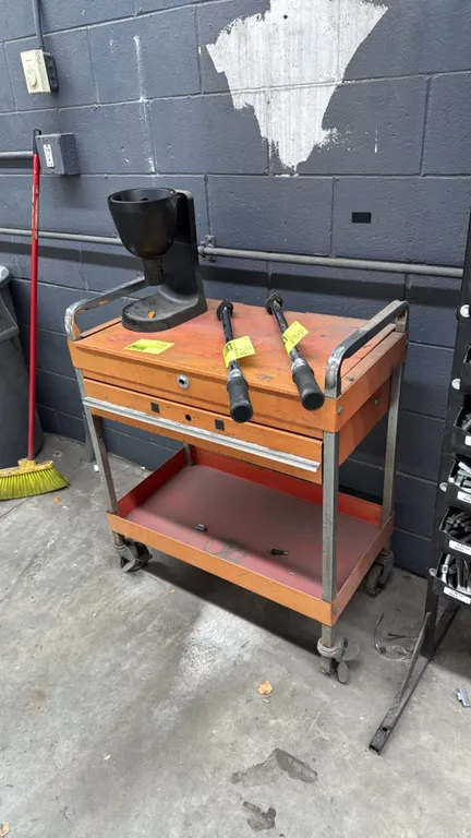 Metal rolling work cart. Features an orange finish, top work surface with dual side handles, pull-out drawer, and open lower shelf. 30"W. Mounted on c
