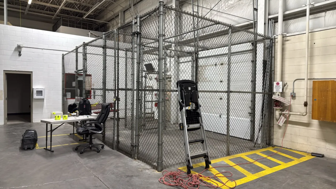 Chain-link security cage. Features galvanized steel construction, wire mesh ceiling panels, and hinged access door with emergency exit panic push bar
