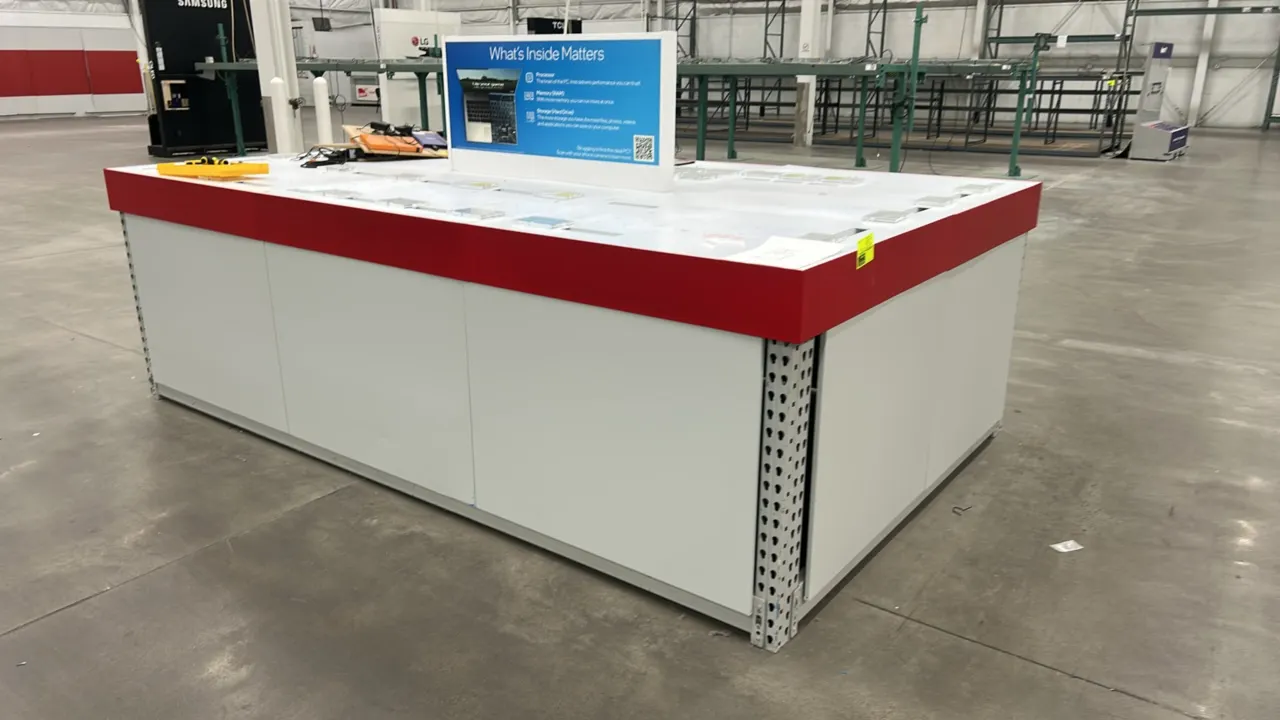 Retail island merchandiser display table. Built on a metal teardrop pallet racking frame with white side panels and red perimeter trim. Features a top