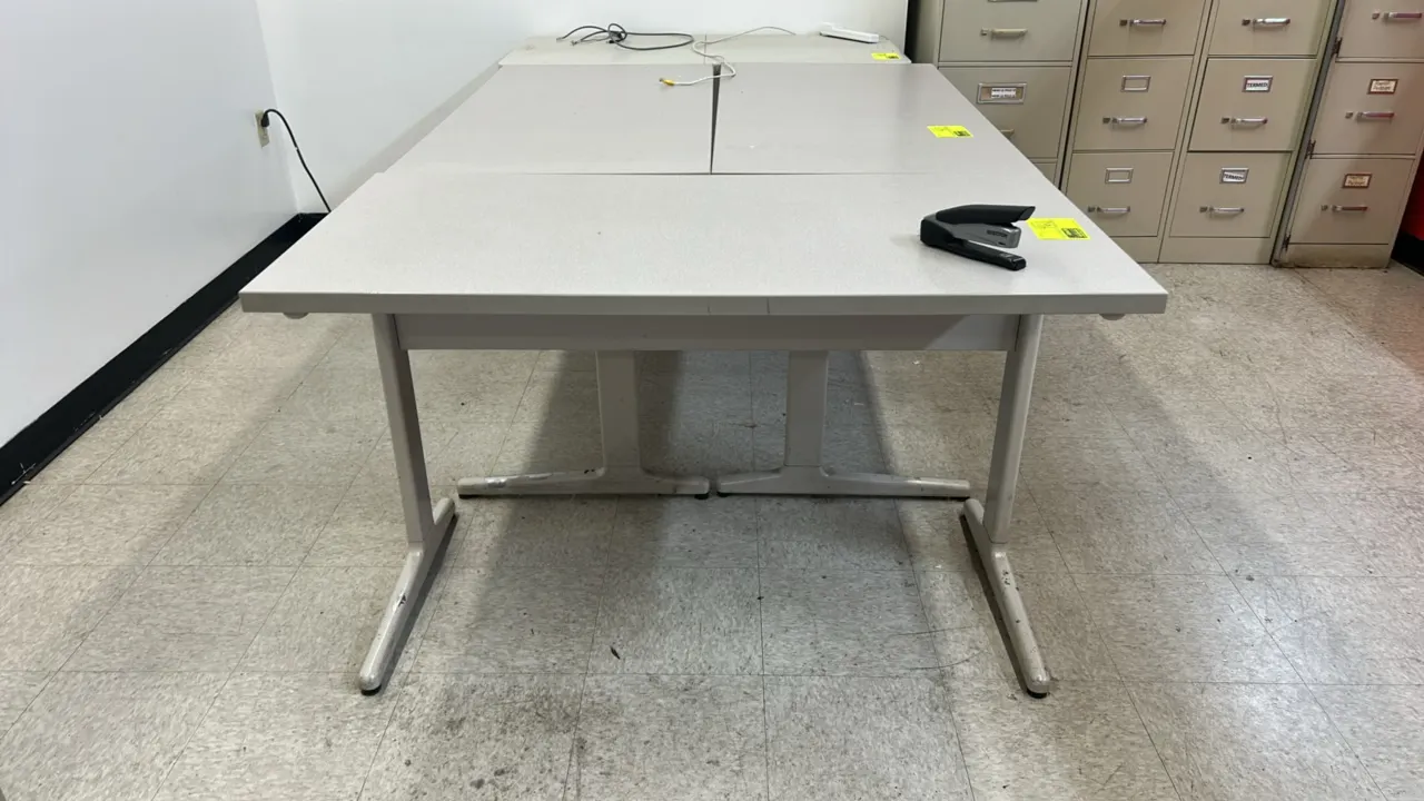 Laminate top utility table. Features a gray laminate work surface. Dimensions: 4' x 2'. Mounted on a metal T-leg base. Note: Stapler and adjacent tabl