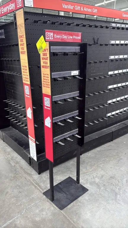 Freestanding pegboard merchandise display rack. Features black metal construction, pegboard back panel, hanging display hooks with integrated clear la