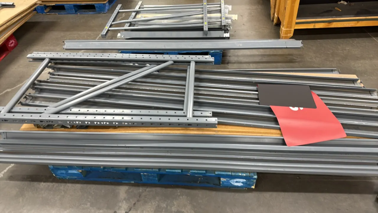 Light-duty racking components. Includes gray metal upright frames, horizontal shelf beams, and wood decking panels. All items stacked on wooden pallet