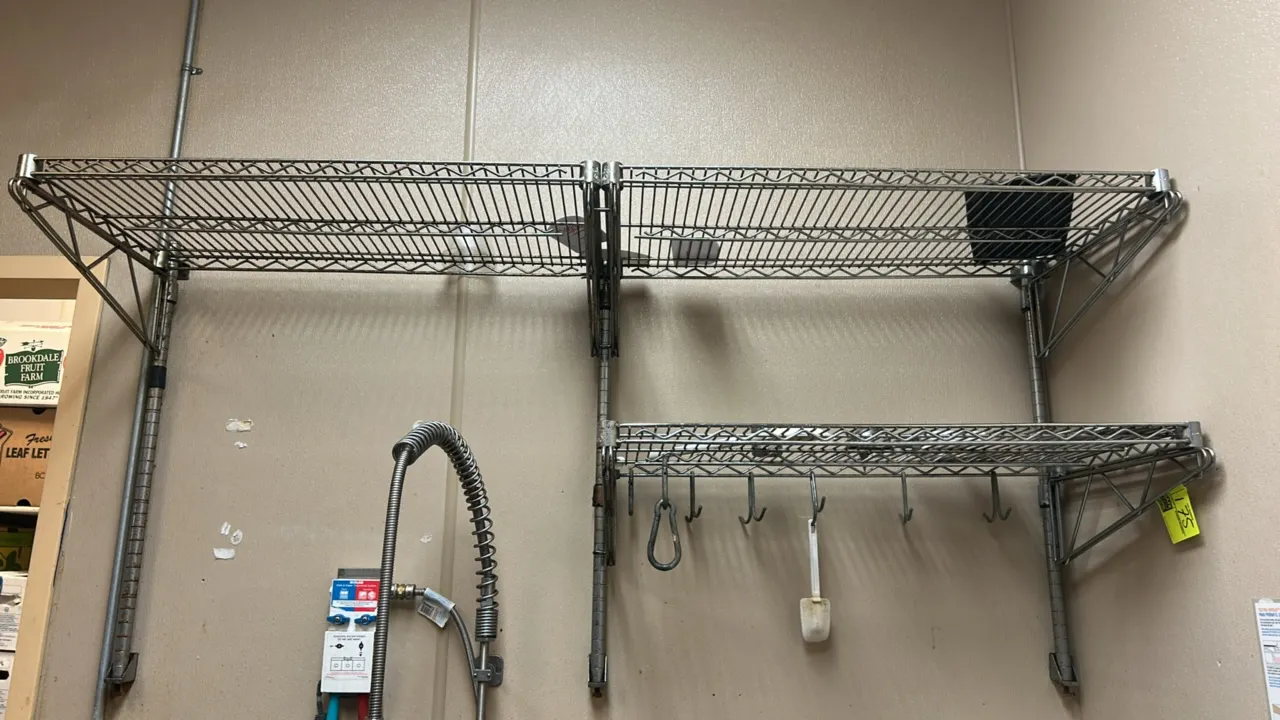Metal wire overhead 2-tier shelving unit. Features wire grid shelves, wall-mounting uprights with support brackets, and hanging hooks along the lower