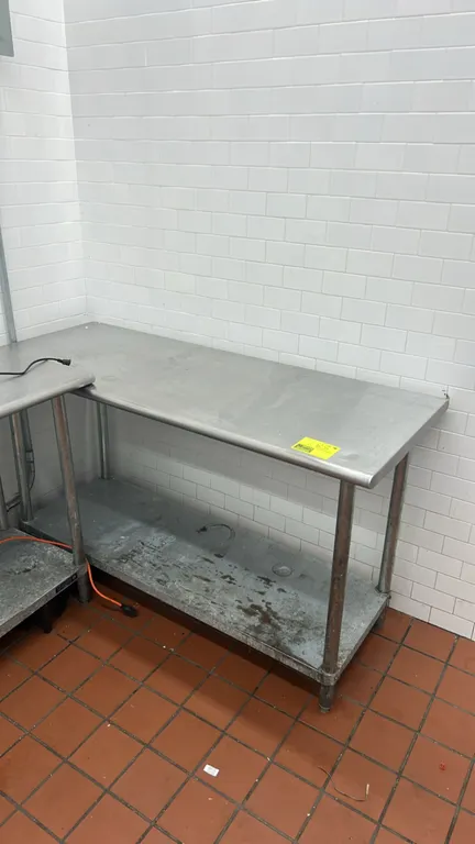Stainless steel work table. Features a flat top with rolled front edge and lower undershelf. Dimensions: 5' x 2'. Mounted on tubular legs with adjusta