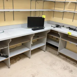 L-shaped laminate countertop workstation with wall-mounted shelving. Features under-counter storage compartments and adjustable wall-mounted shelf sta